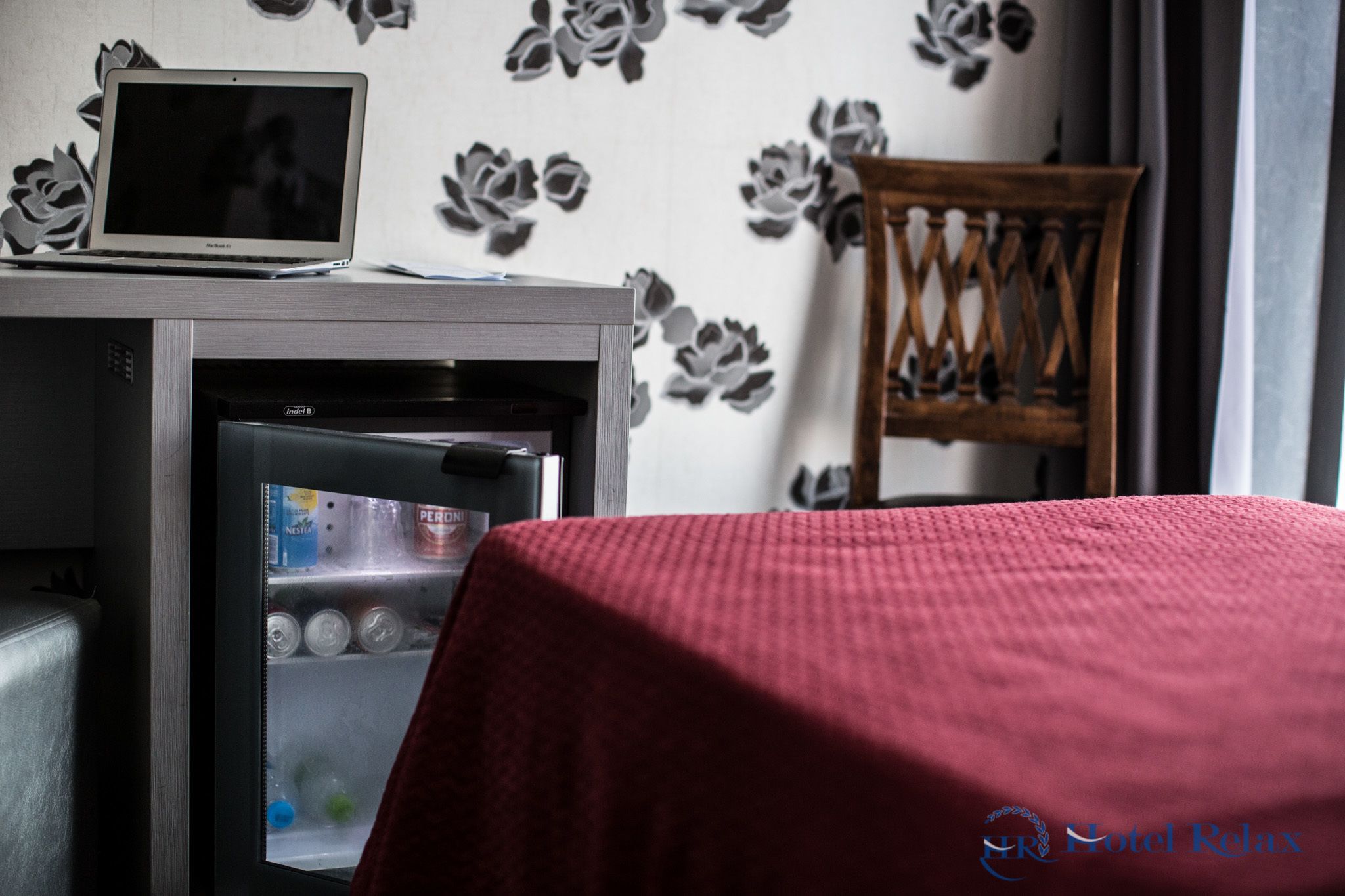 A hotel room with a desk, a laptop, a mini-fridge, and a red bedspread.