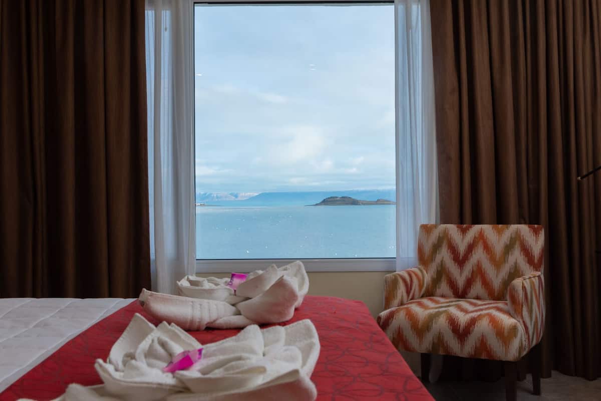 A hotel room with a bed, a chair, and a window with a view of the sea and mountains.