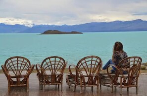 A person sitting on wicker chairs facing a calm lake with mountains in the background.