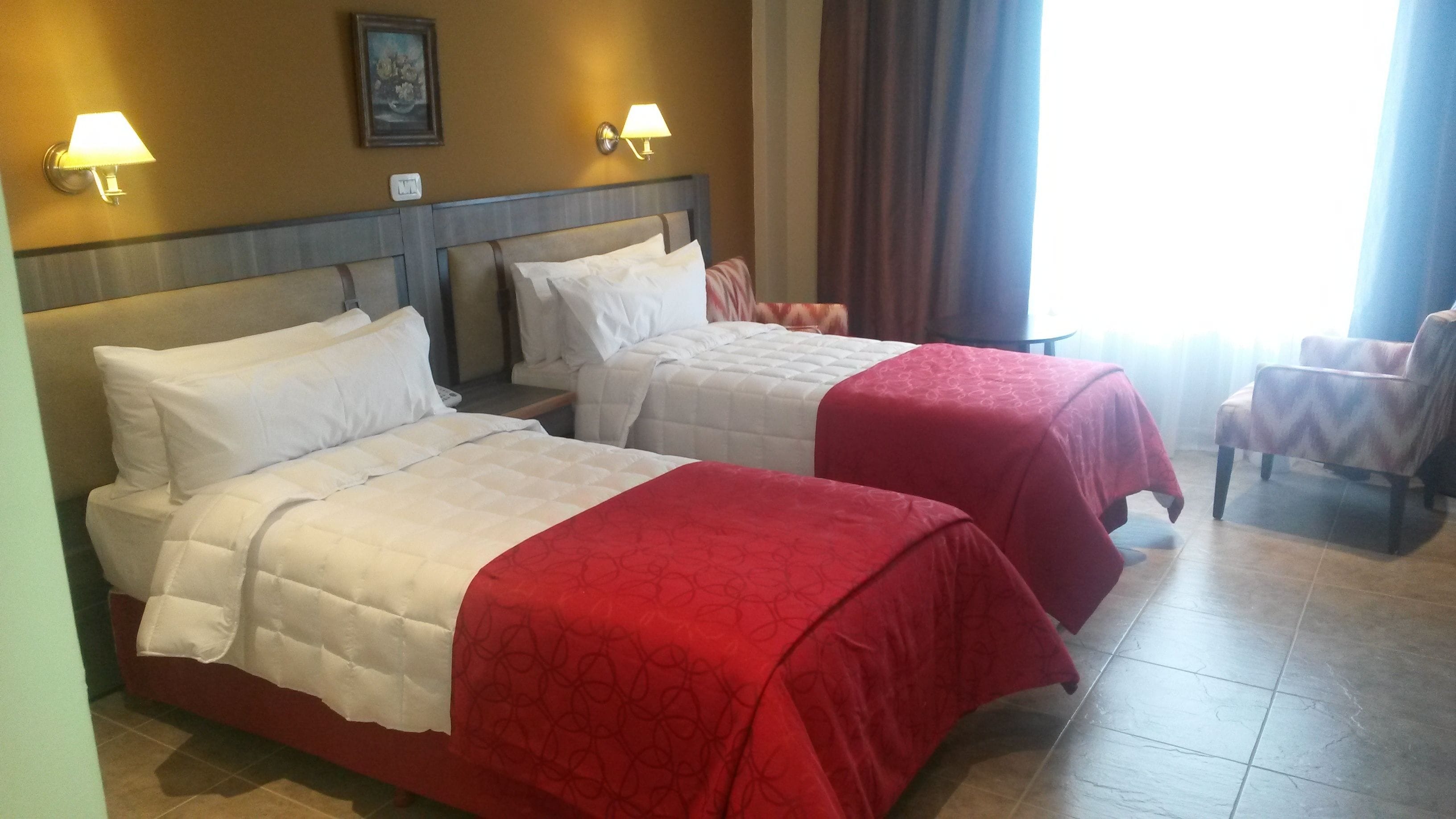 Dos camas con ropa de cama roja y blanca, un cabecero de madera, un cuadro en la pared y una ventana con cortinas.