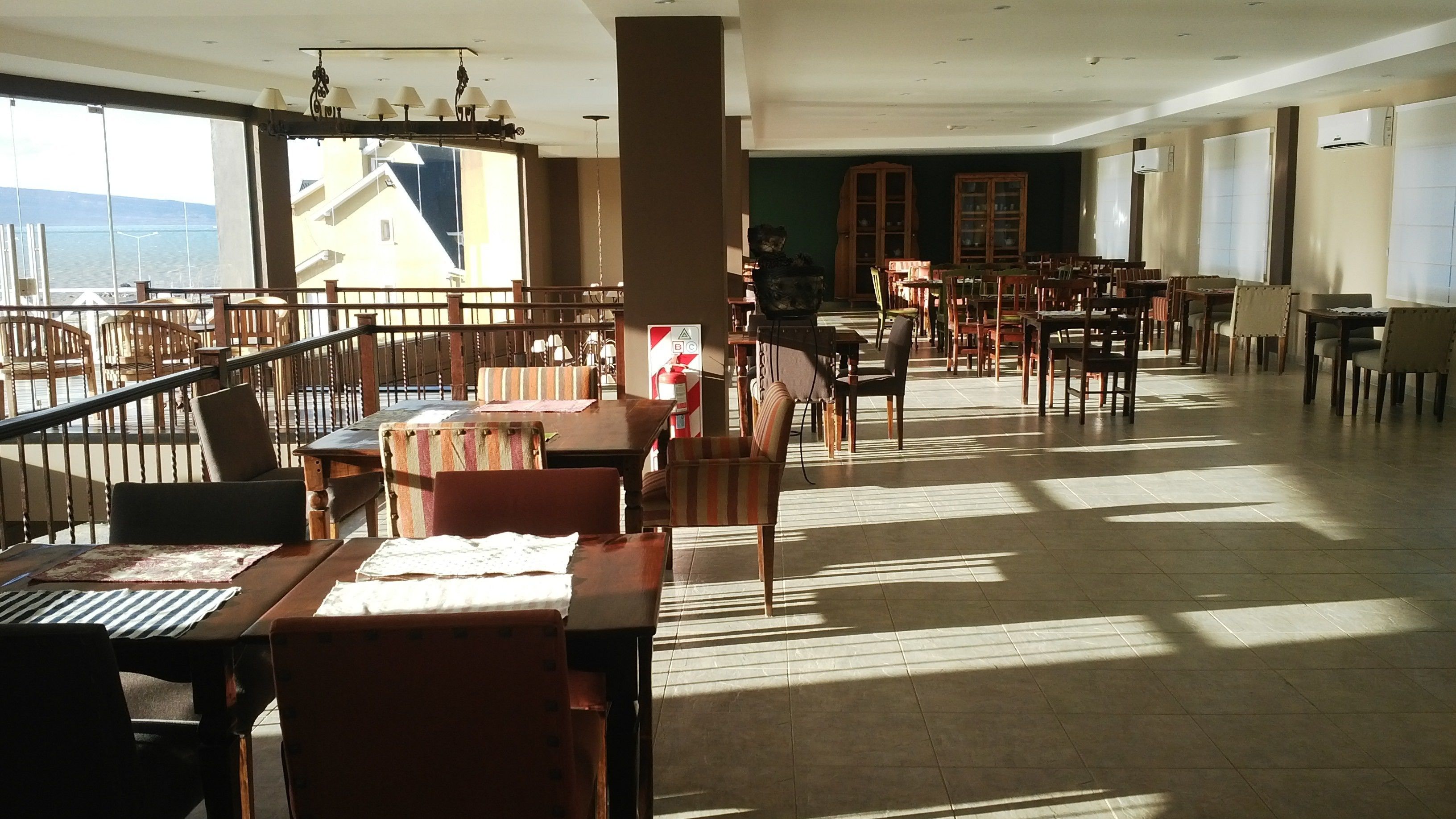 A spacious indoor area with multiple wooden tables and chairs, large windows offering views of buildings and a body of water, and a staircase leading to an upper level.