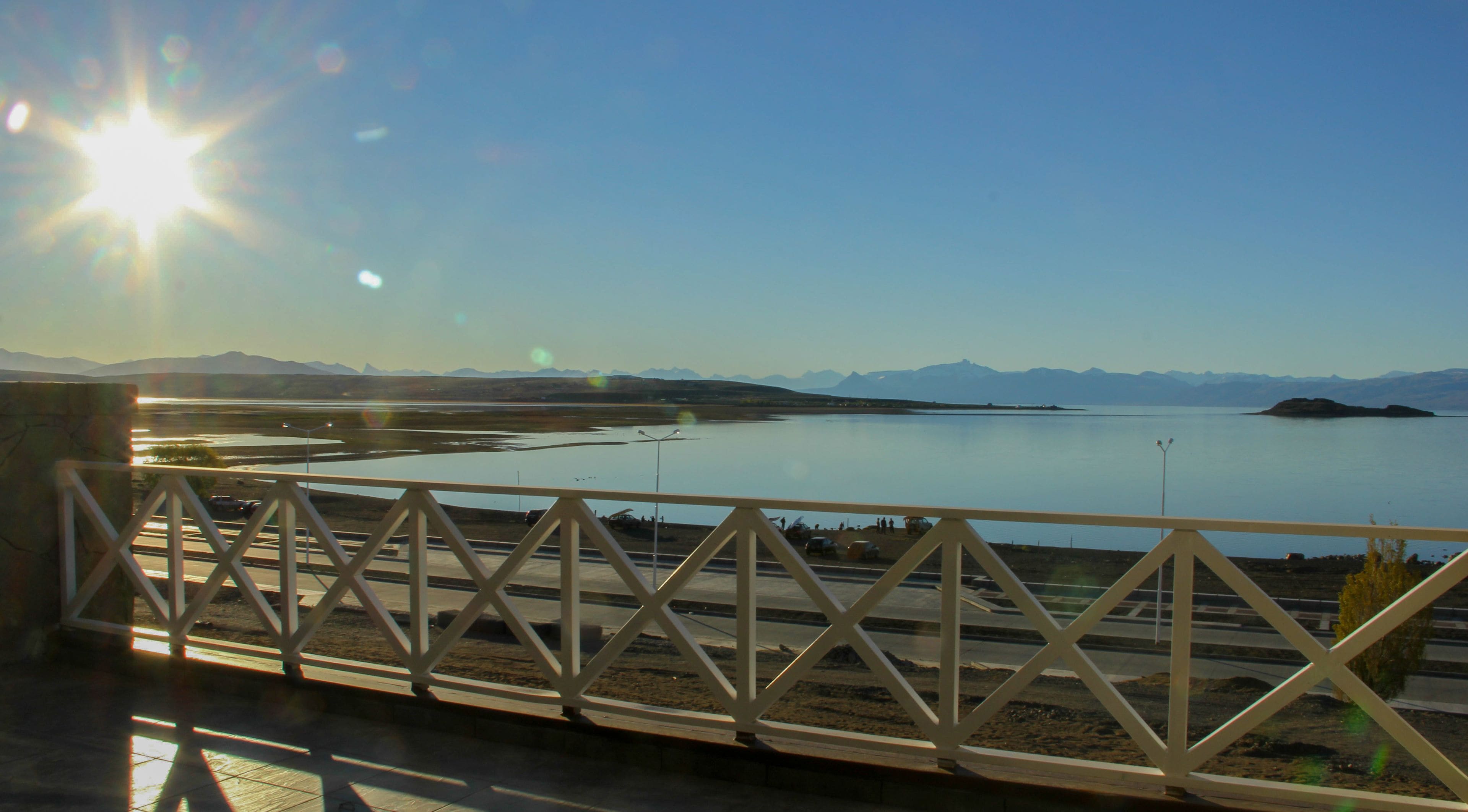 Une vue depuis un balcon donnant sur un lac paisible, avec des montagnes lointaines, un ciel dégagé et une rambarde en métal.