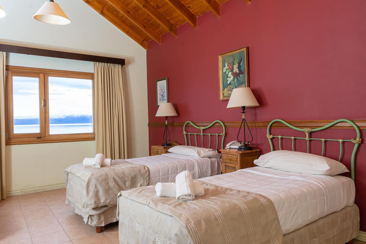 A hotel room with two beds, a large window with curtains, wooden ceiling beams, and a painting on the wall.