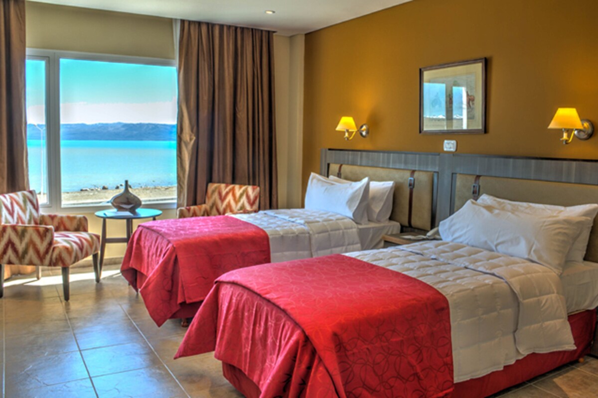 A hotel room with two beds, a view of the beach, a table with a lamp, and a chair.