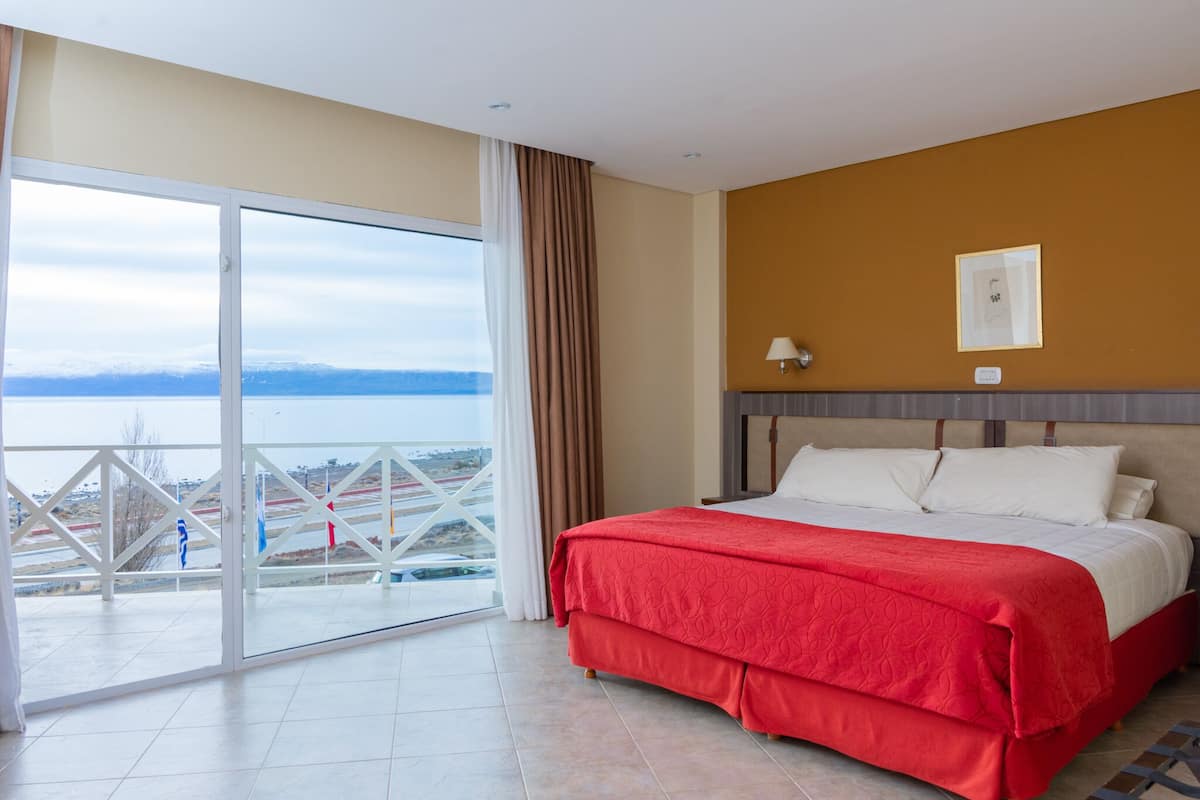 A hotel room with a large sliding glass door leading to a balcony with a view of a body of water and mountains. The room has a bed with a red and white bedspread, a headboard, and a wall-mounted lamp.