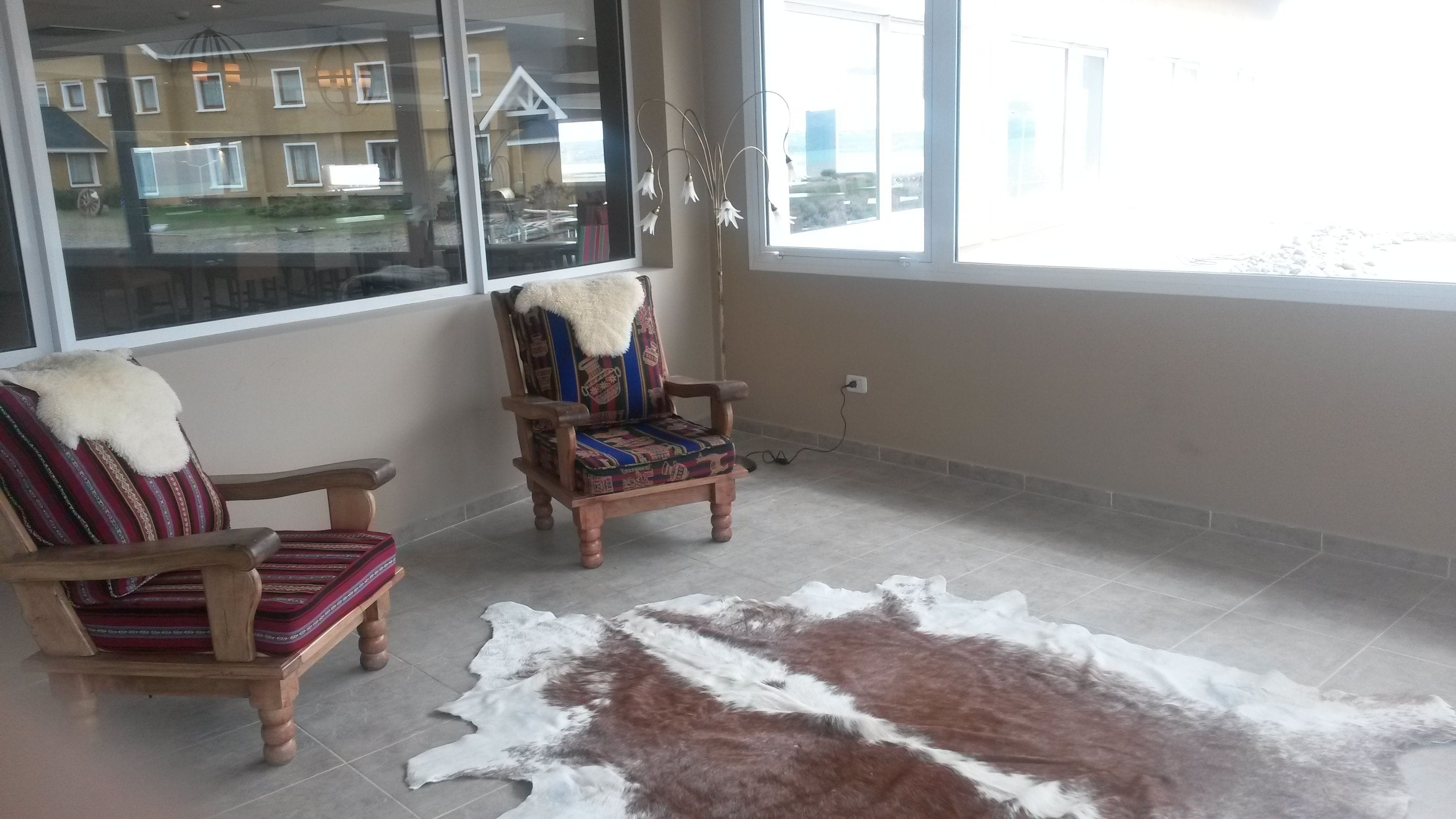 A room with two wooden chairs with patterned cushions, a large fur rug, and a view of a residential area through a large window.
