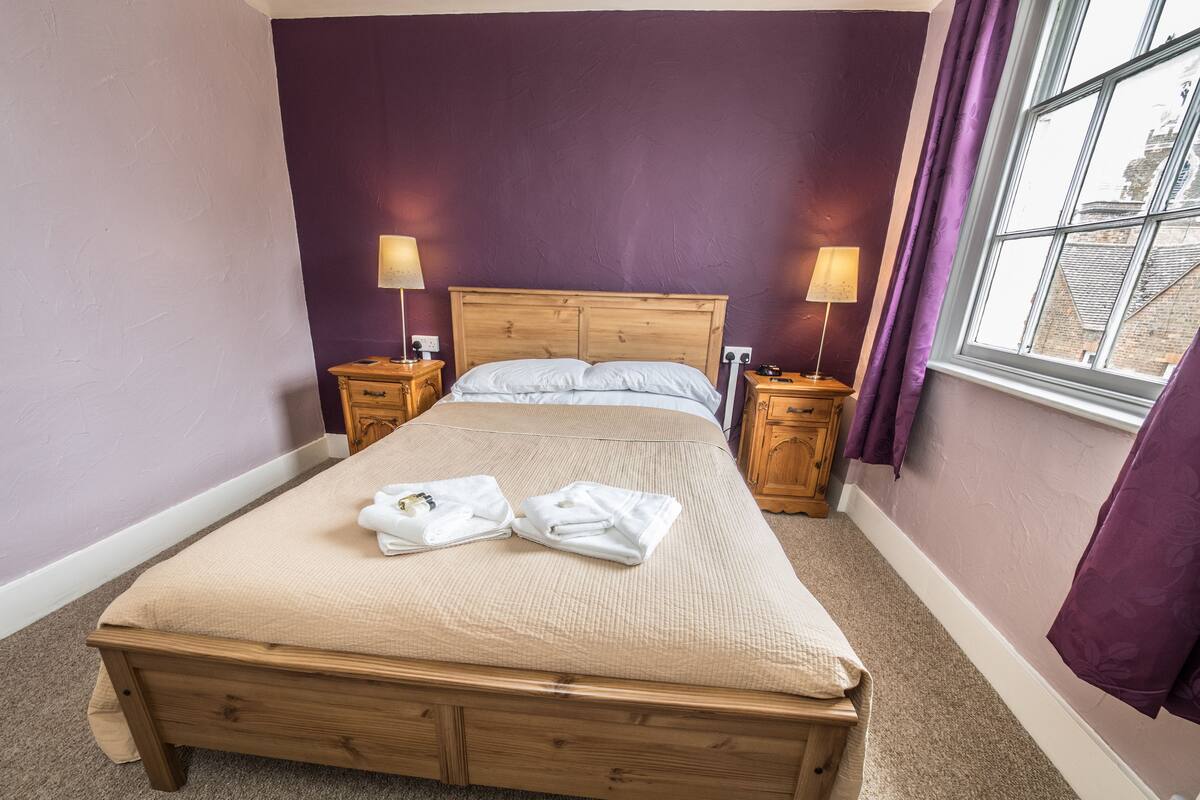 A neatly made bed with a beige bedspread, two wooden nightstands with lamps, and a window with purple curtains.