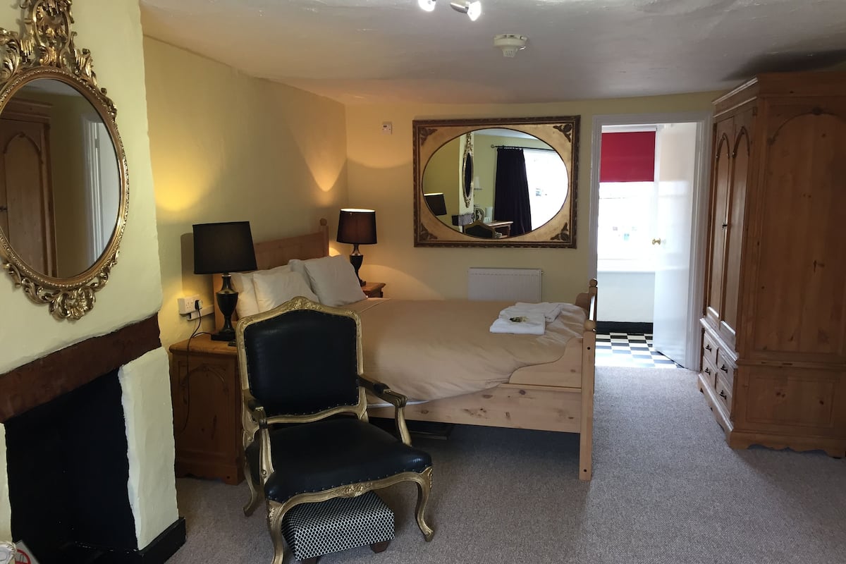 A bedroom with a bed, a chair, a mirror, a dresser, and a fireplace.