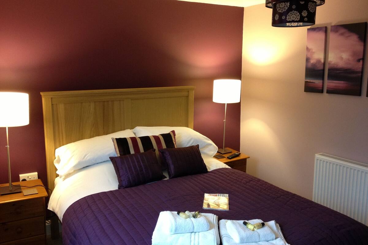 A hotel room with a bed, two bedside lamps, a nightstand, a dresser, and a wall with two framed pictures.