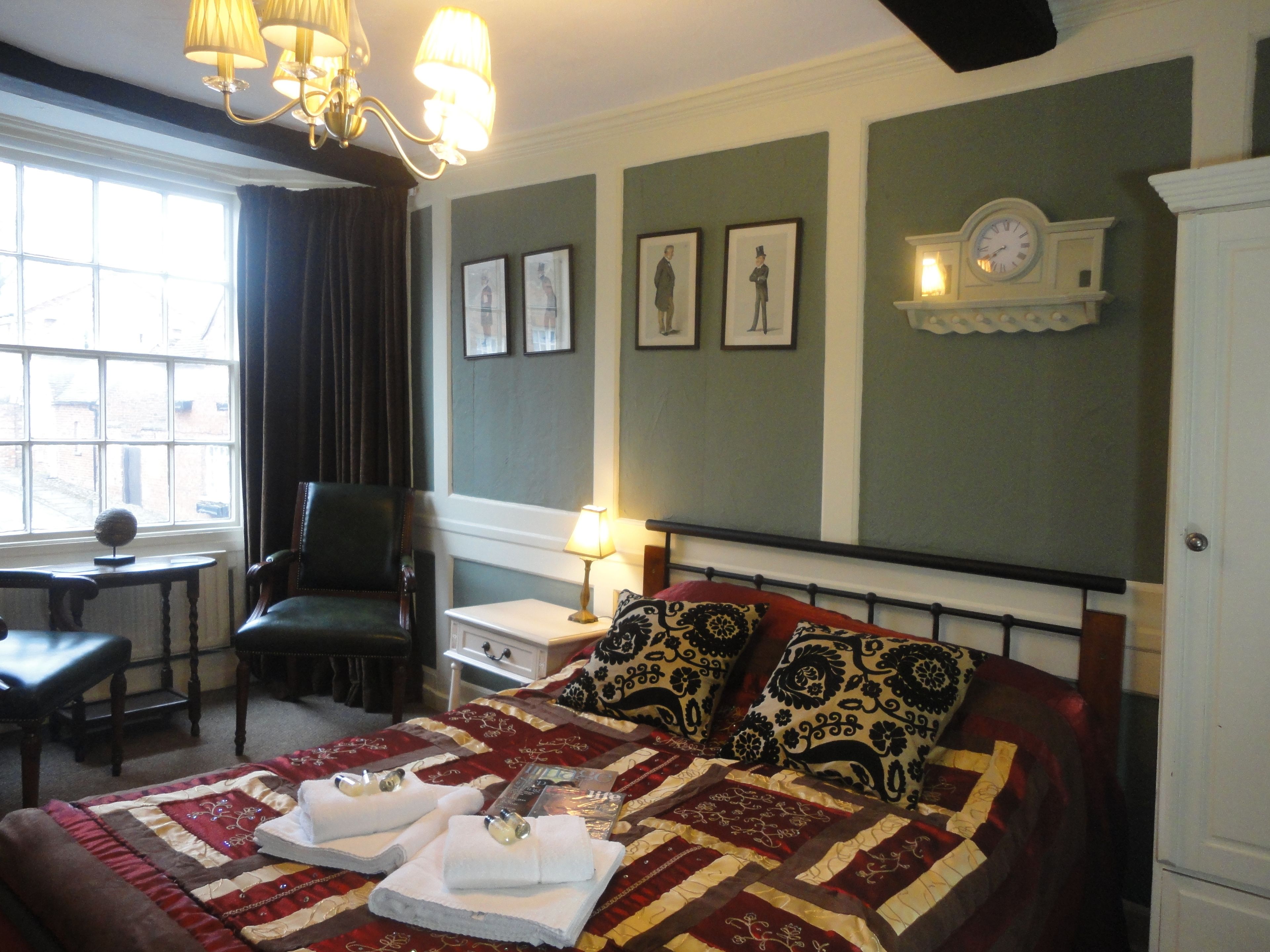 A hotel room with a bed, a desk, a chair, a lamp, and a clock on the wall.