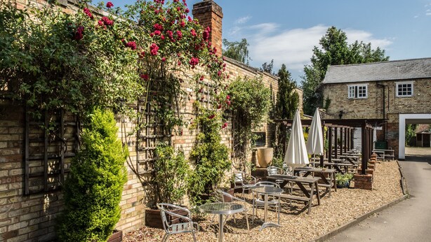 A brick building with a garden patio featuring tables and chairs, a pergola with climbing plants, and a chimney. - Longs Inn (Milton Keynes)
