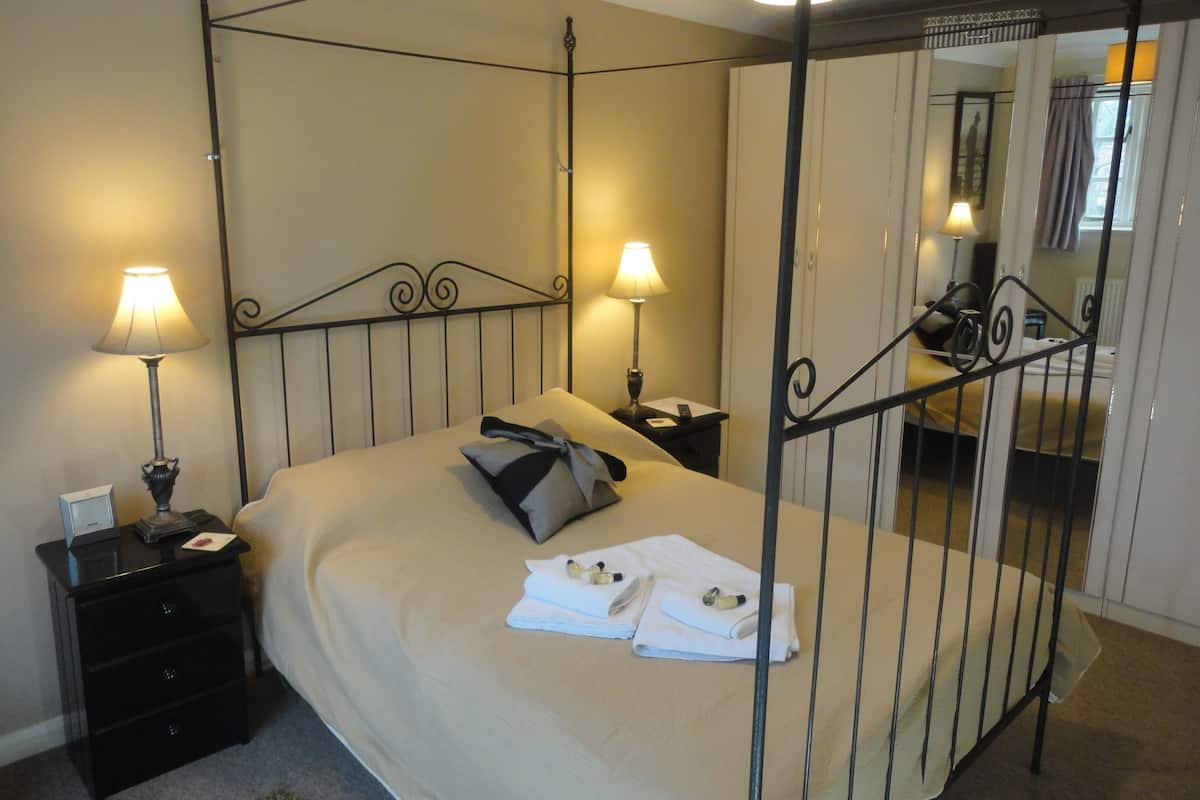 A four-poster bed with a beige bedspread and pillows, two bedside tables with lamps, a mirror, and a view of a window with curtains.