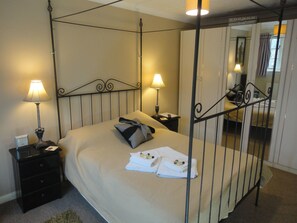 A four-poster bed with a beige bedspread and pillows, two bedside tables with lamps, a mirror, and a view of a window with curtains.