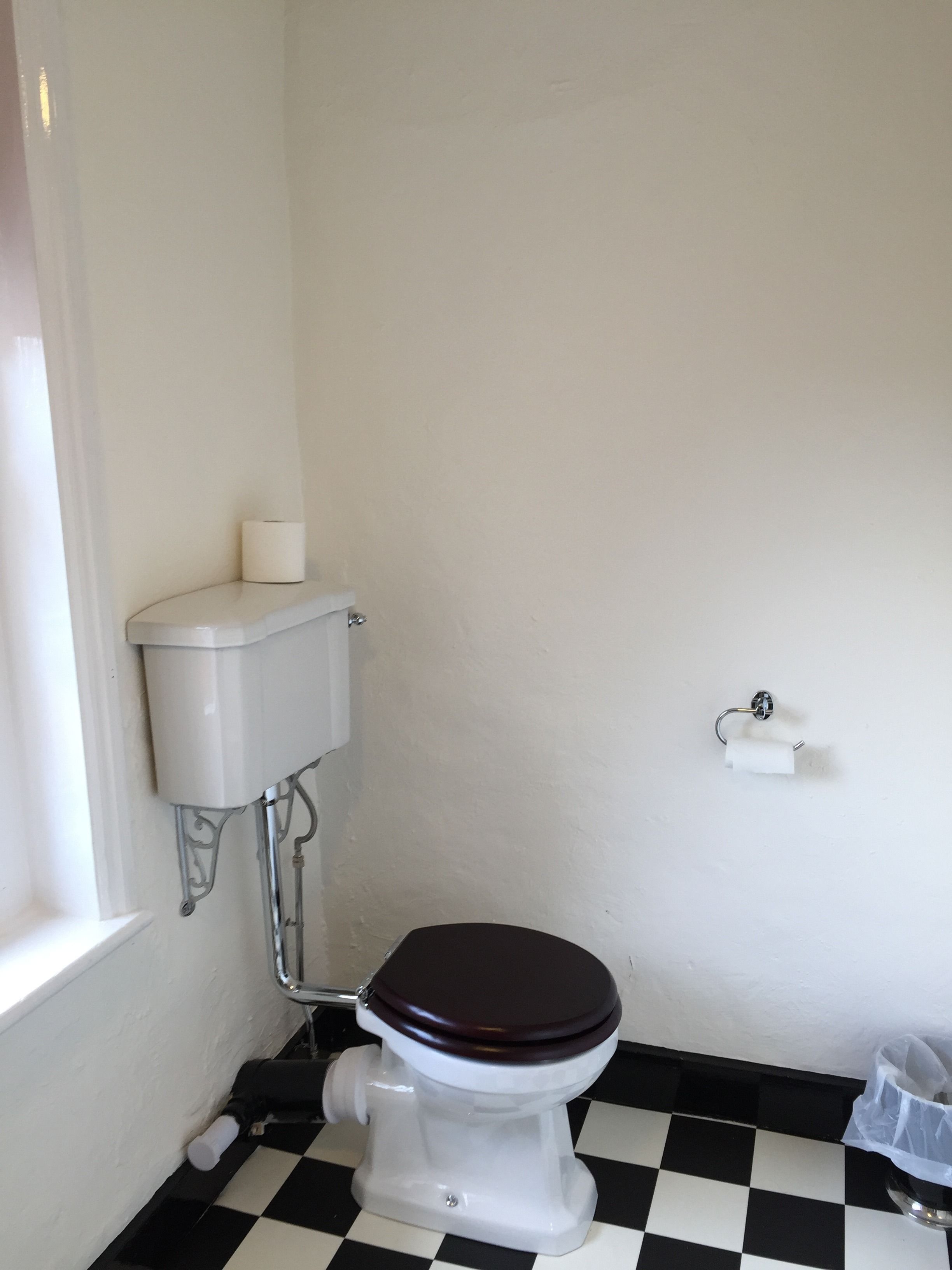Un inodoro pequeño y blanco con una tapa marrón, un dispensador de papel higiénico blanco en la pared y un suelo a cuadros blanco y negro.