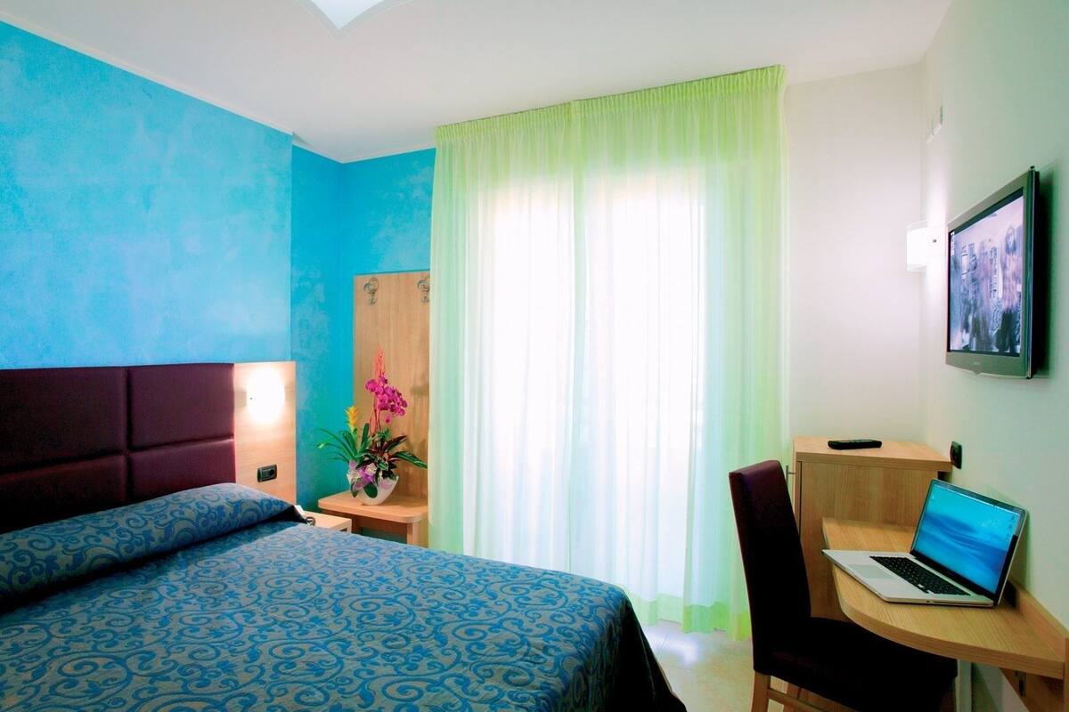 Una habitación de hotel con cama, escritorio con computadora portátil, silla, televisor y ventana con cortinas.