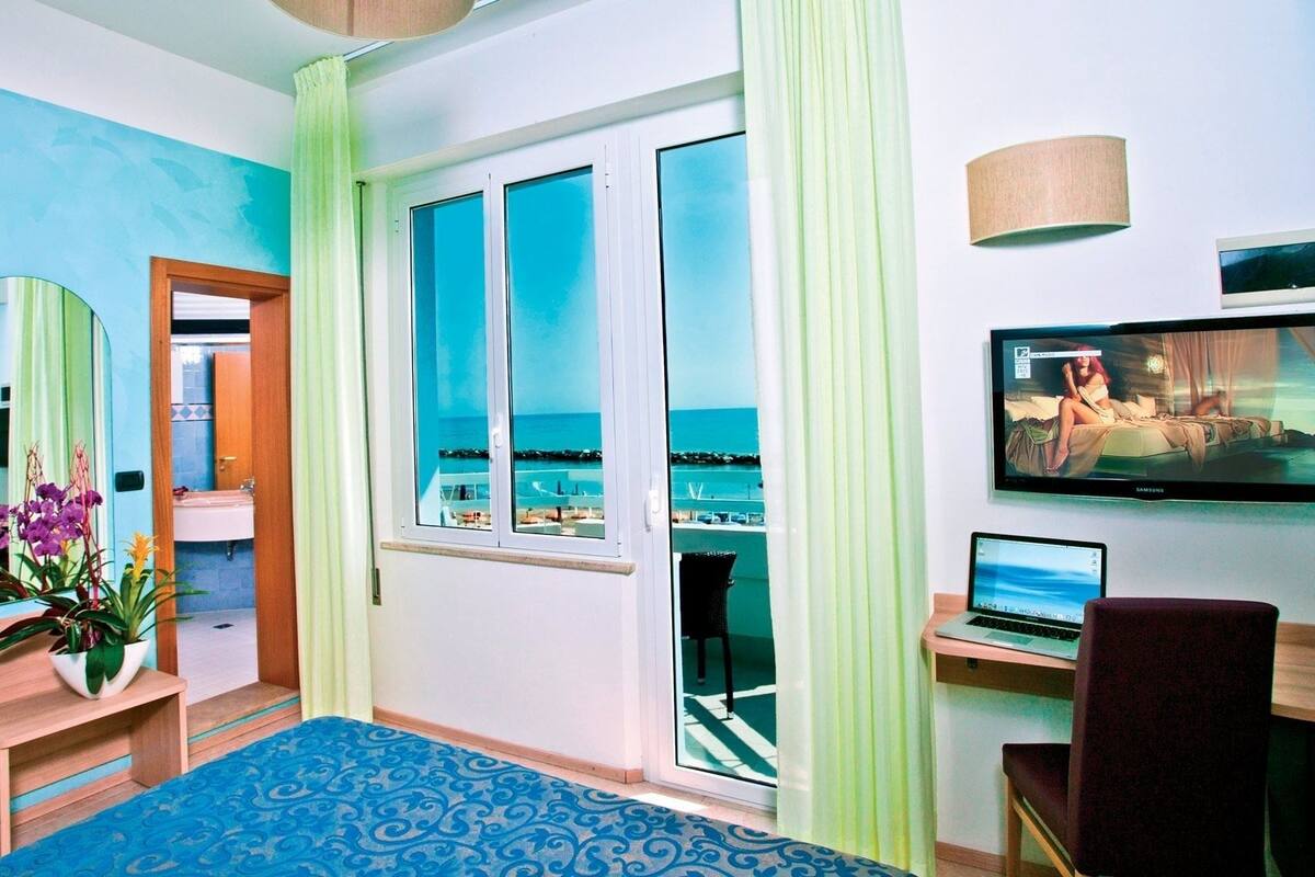 Habitación de hotel con un ventanal grande, una cama, un escritorio con una computadora portátil, un televisor y un espejo.