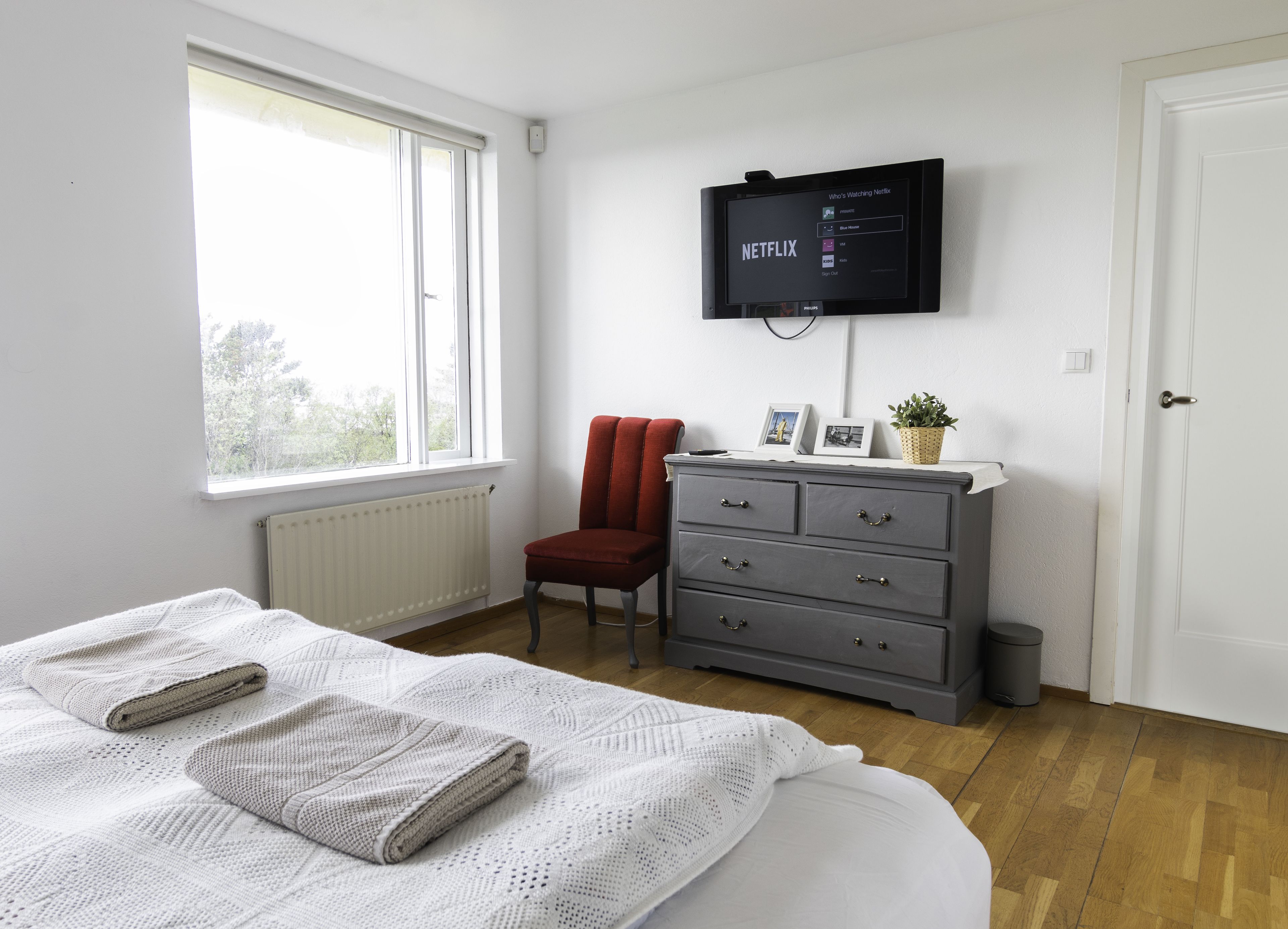 A bedroom with a bed, a dresser, a chair, a TV mounted on the wall, and a window.