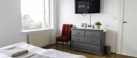 A bedroom with a bed, a dresser, a chair, a TV mounted on the wall, and a window.