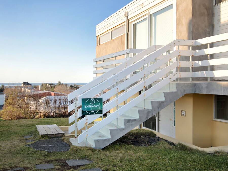 A building with a concrete exterior, a white staircase with a sign indicating an entrance, and a view of the surrounding area.