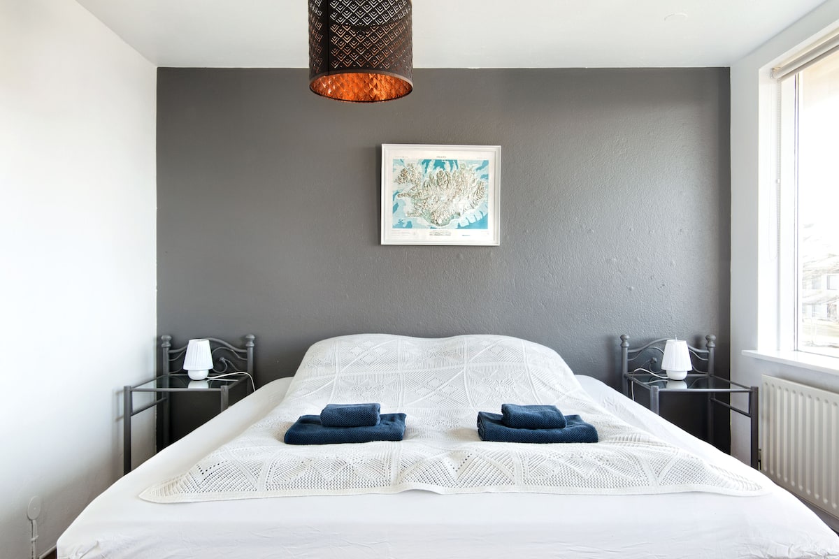 Un hotel moderno con una cama grande, dos mesitas de noche con lámparas, un mapa enmarcado en la pared y vistas de la ciudad a través de la ventana.