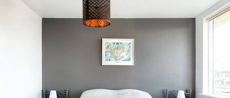 Un hotel moderno con una cama grande, dos mesitas de noche con lámparas, un mapa enmarcado en la pared y vistas de la ciudad a través de la ventana.