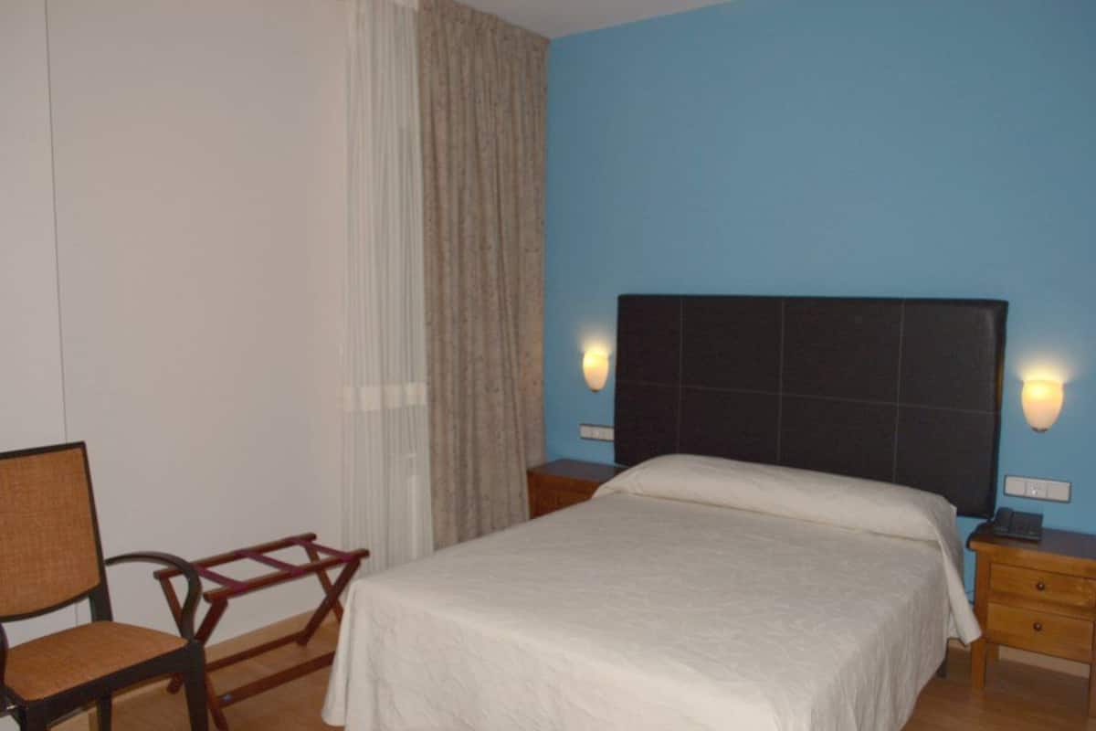 Habitació d'hotel amb un llit, una cadira, una taula de nit i una làmpada penjada a la paret.