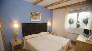 Un dormitorio con dos camas, cabeceros de madera, una mesita de noche con una lámpara, un televisor sobre un mueble de madera y una ventana con cortinas.
