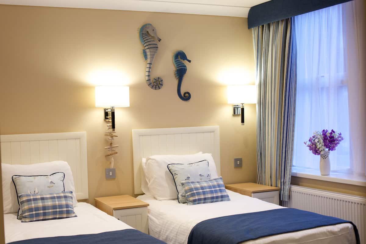 A hotel room with two beds, a seahorse wall decoration, a window with curtains, and a vase with flowers on the windowsill.