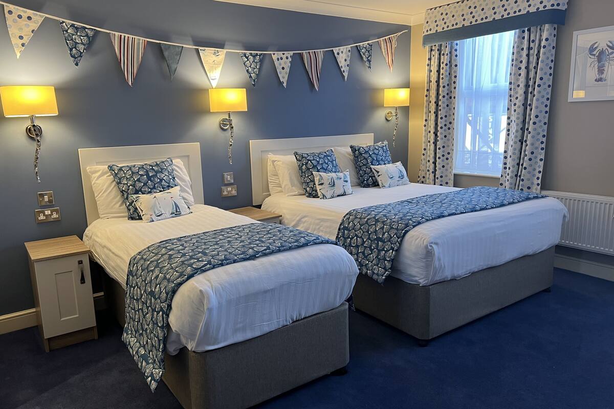 A double room with two beds, each with matching headboards and bedside tables. There are two wall-mounted lamps and a window with curtains.