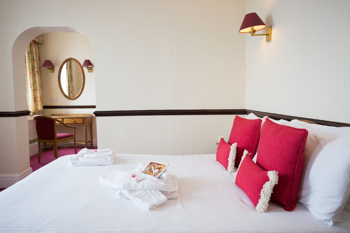 A neatly made bed with red pillows, a desk with a chair, and a mirror.