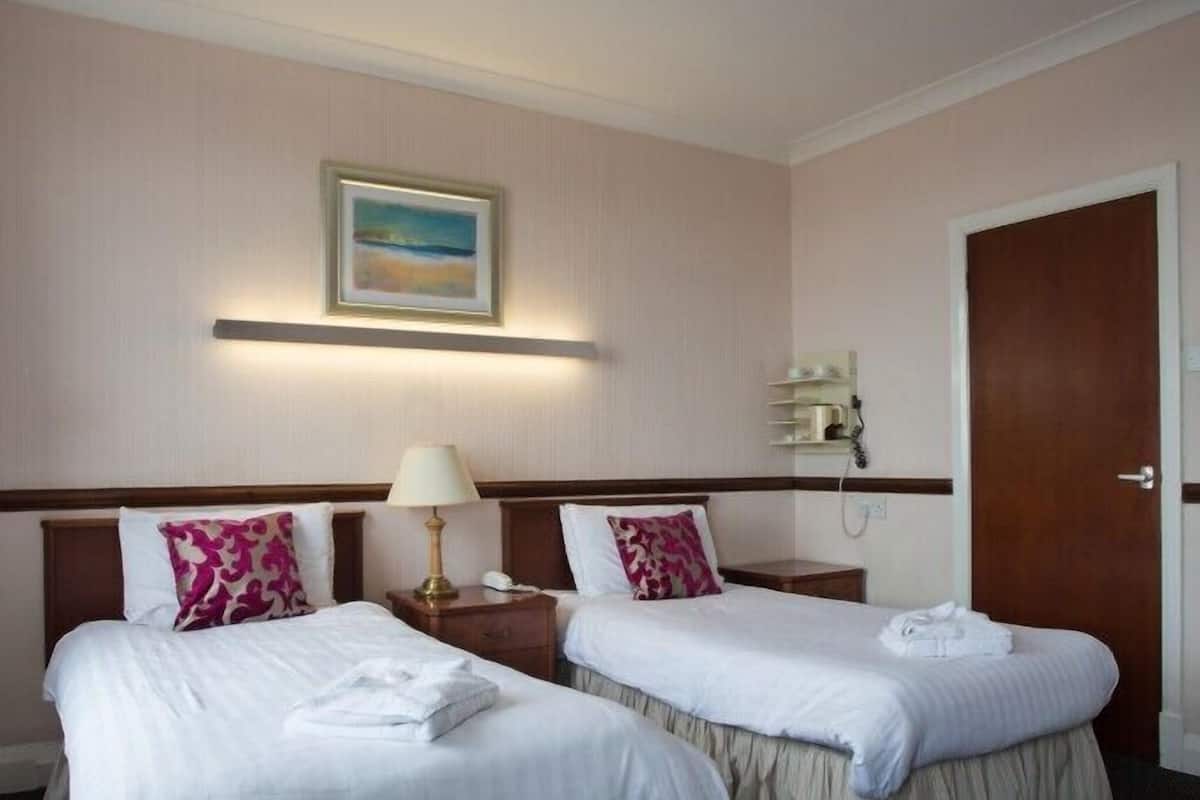 A hotel room with two beds, a nightstand with a lamp, a painting on the wall, and a door.