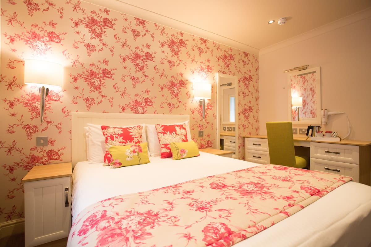 A bedroom with a floral wallpaper, a bed with multiple pillows, bedside lamps, a desk with a chair, and a mirror.