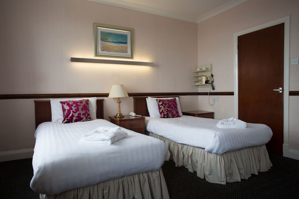 A hotel room with two beds, a nightstand with a lamp, a painting on the wall, and a door.
