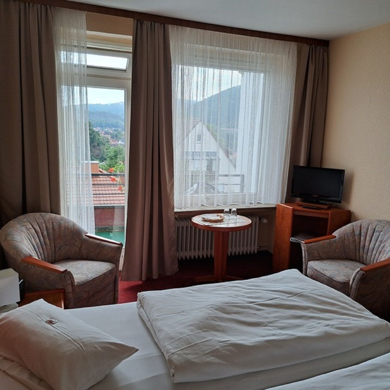A hotel room with two armchairs, a bed, a small table with a glass, and a TV.