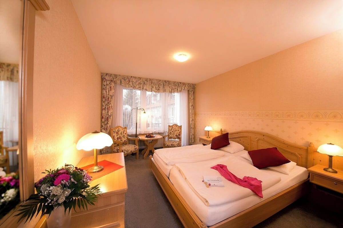 Habitación de hotel con una cama grande, dos mesitas de noche con lámparas, una pequeña mesa con sillas y una ventana con cortinas.