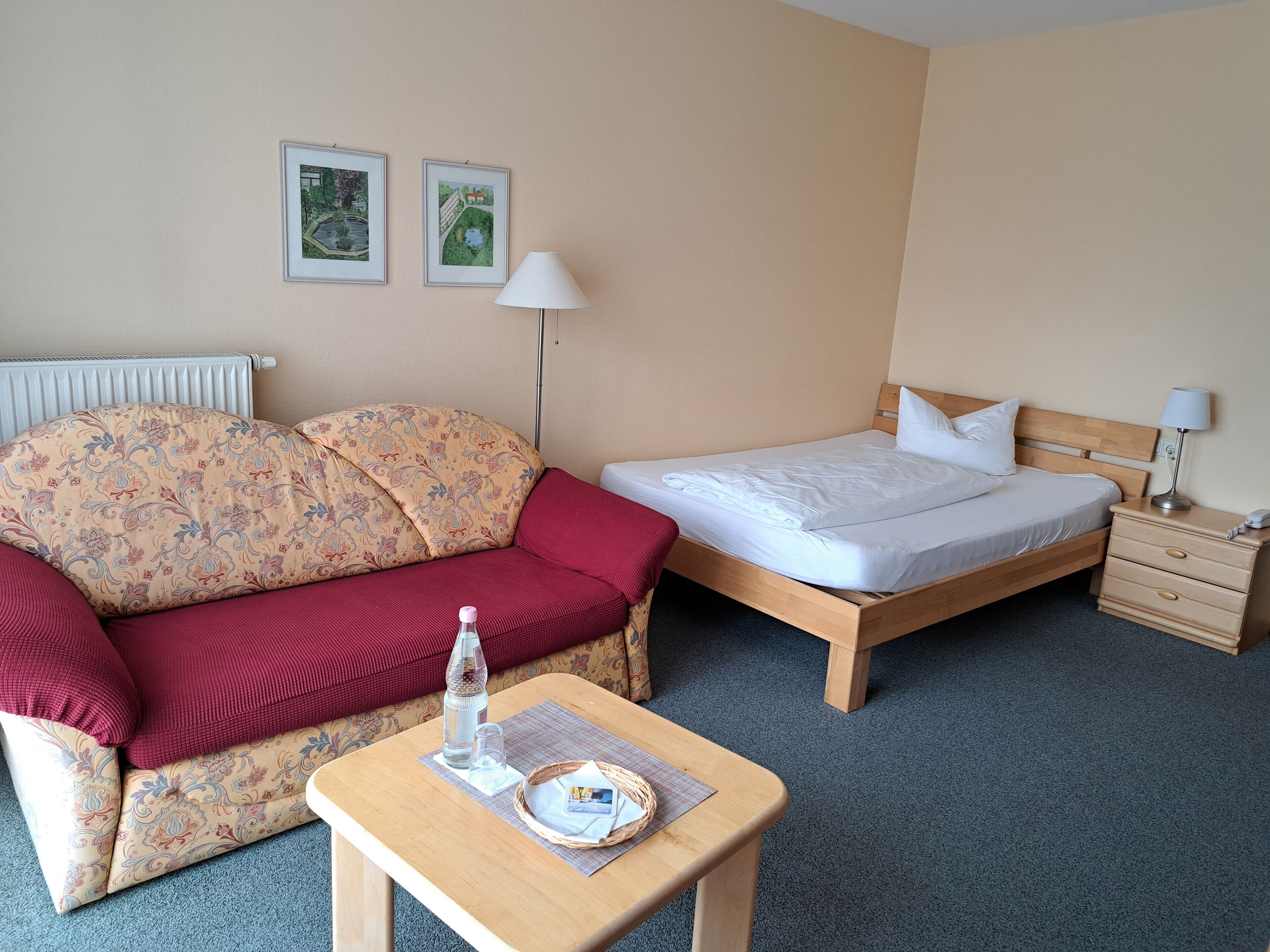 Ein Hotelzimmer mit einem Bett, einem Sofa, einem Tisch mit Flasche und Gläsern sowie zwei gerahmten Bildern an der Wand.