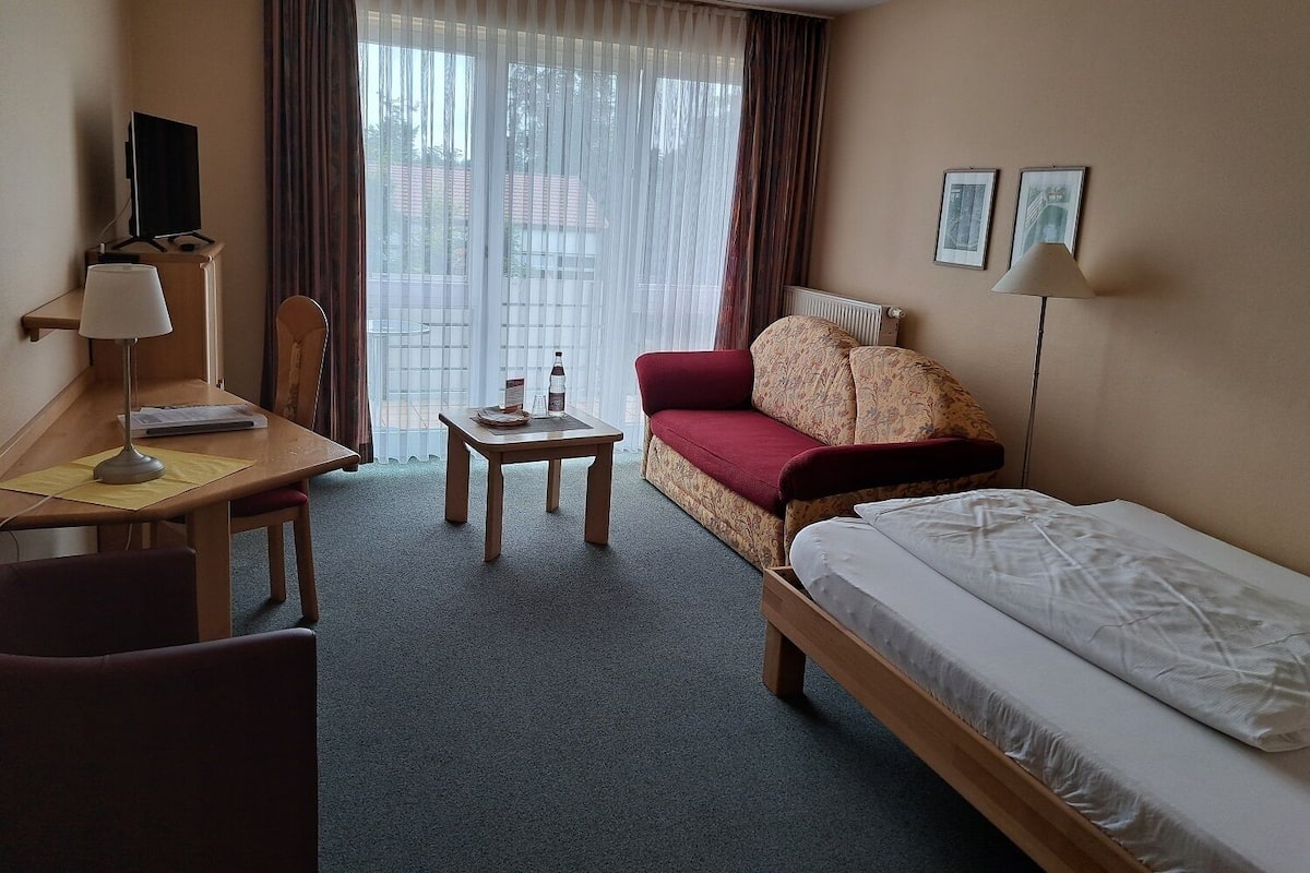 Habitación de hotel con cama, escritorio, silla, mesita pequeña, sofá y ventana con cortinas.