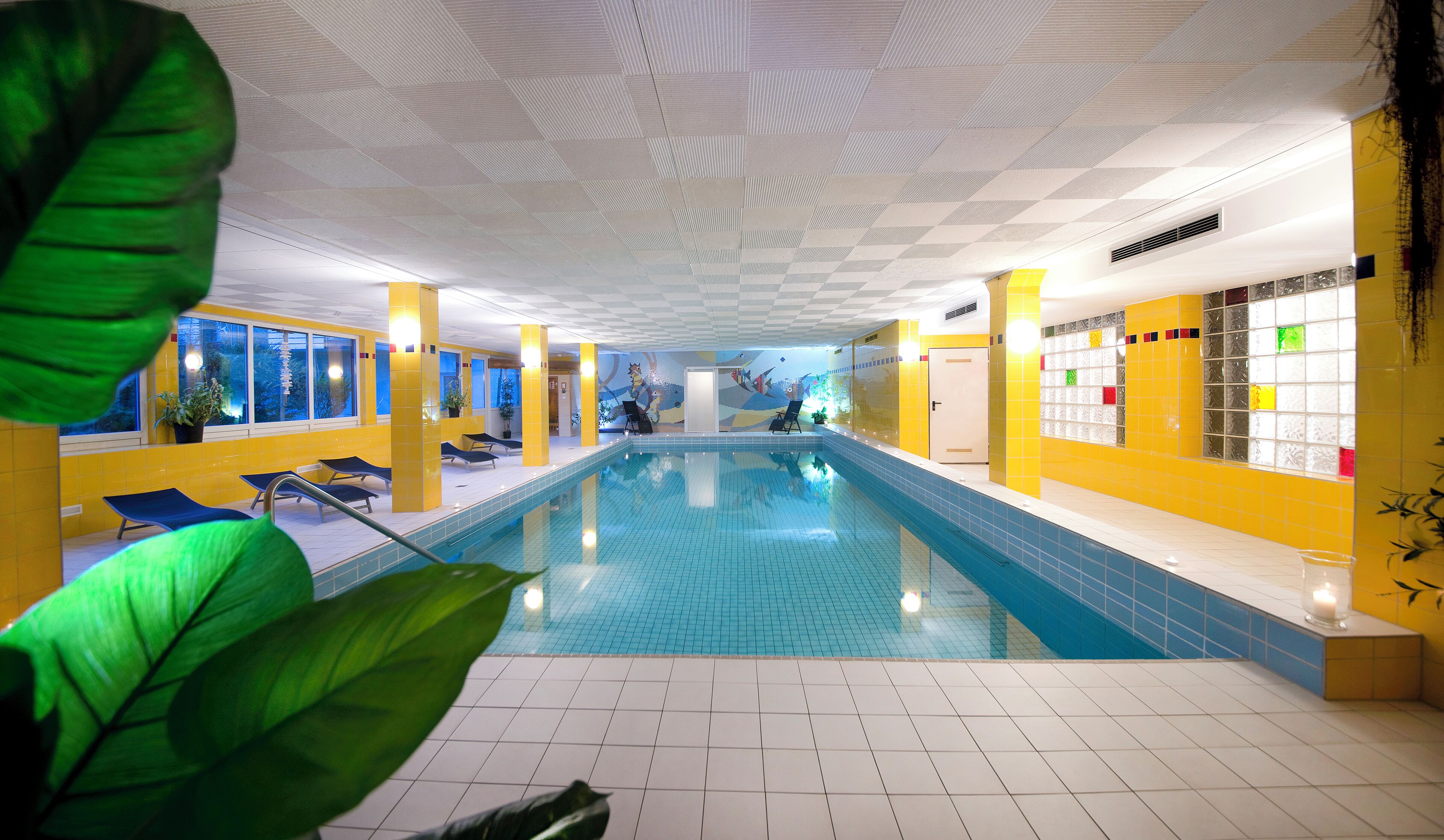 An indoor swimming pool with blue tiles, yellow walls, and a ceiling with a geometric pattern.