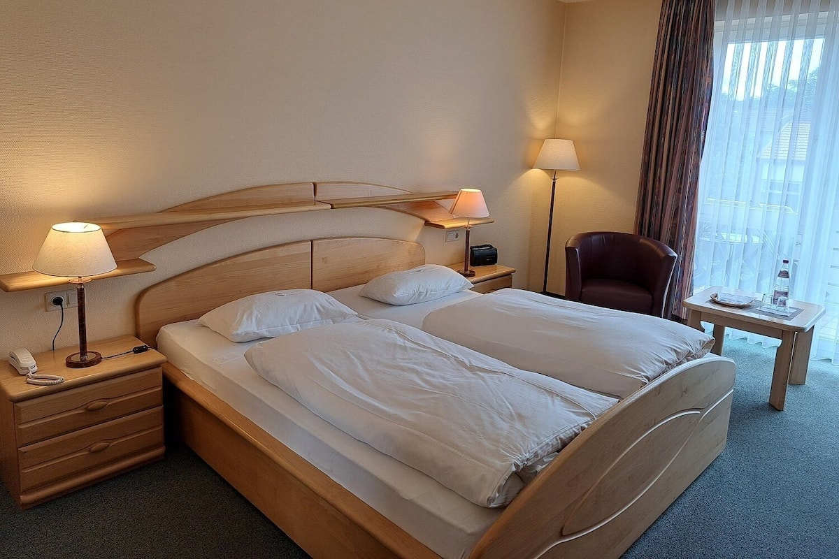 Habitación de hotel con cama de madera, dos mesitas de noche con lámparas, una silla y una pequeña mesa con una botella y un vaso.