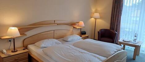 Habitación de hotel con cama de madera, dos mesitas de noche con lámparas, una silla y una pequeña mesa con una botella y un vaso.