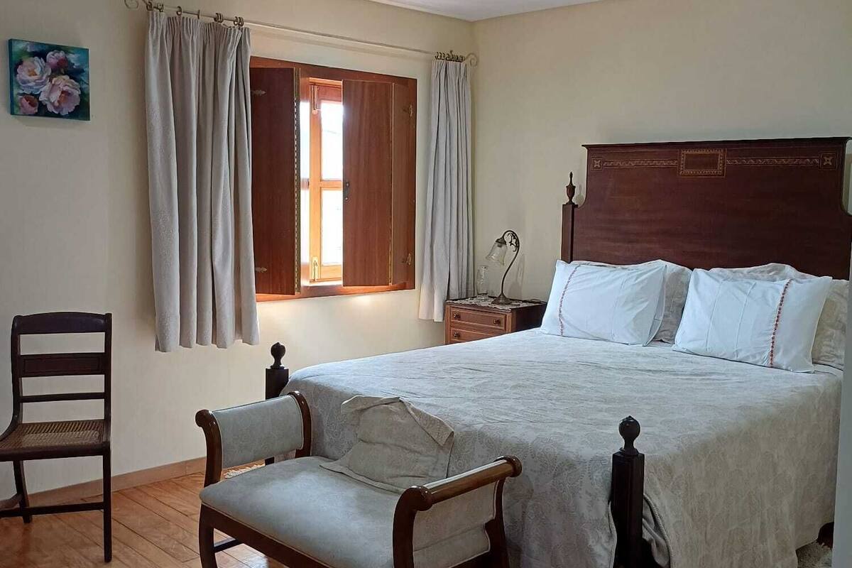 A bedroom with a wooden bed, a chair, a nightstand, and a window with curtains.