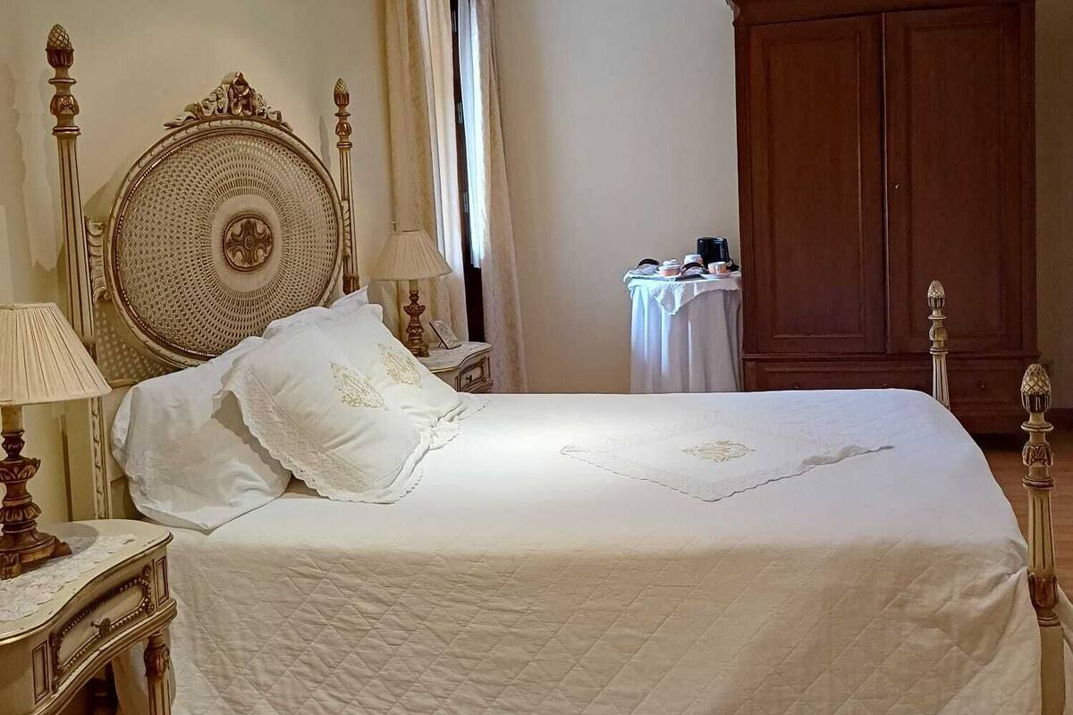 A neatly made bed with a quilted white bedspread, a wooden headboard with intricate carvings, a bedside table with a lamp, and a large wooden wardrobe in a well-lit bedroom.