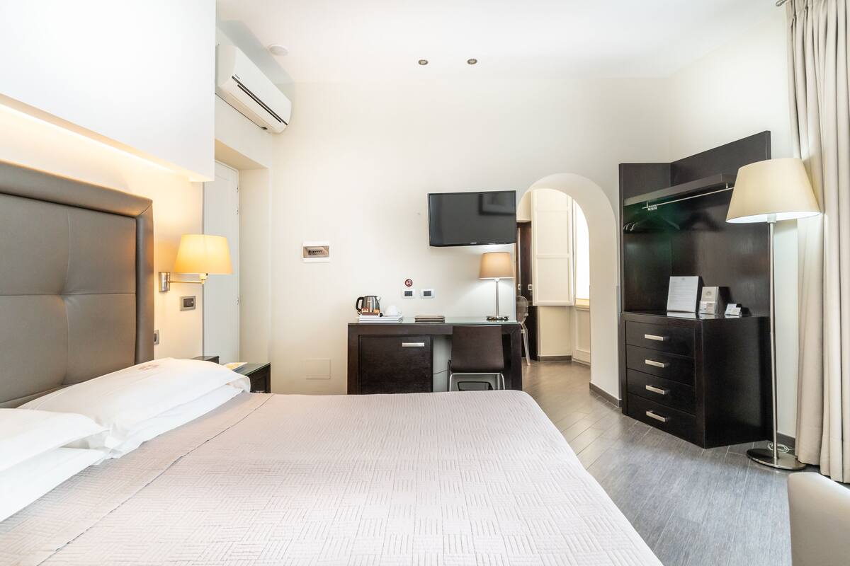 A hotel room with a large bed, a desk with a chair, a wardrobe, a television, and a lamp.