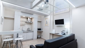 A modern kitchen with a glass countertop, white cabinets, and a microwave. There are two bar stools and a black sofa in the living area. - Magi House Relais (Sorrento)