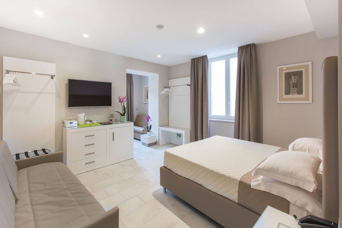 A modern hotel room with a large bed, a sofa, a desk with a flat-screen TV, a mirror, and a window with curtains.
