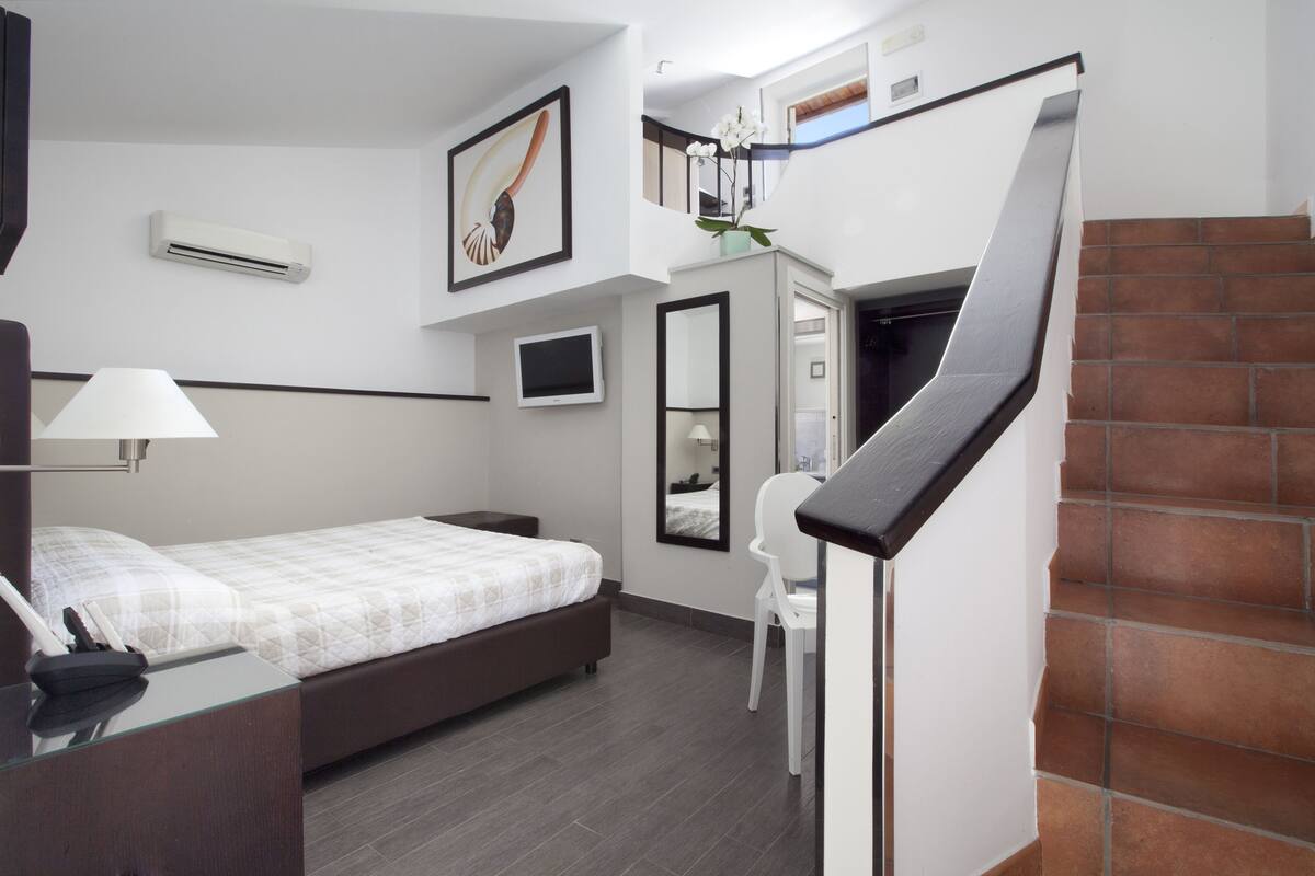 A modern hotel room with a bed, a desk, a chair, a TV, and a staircase.