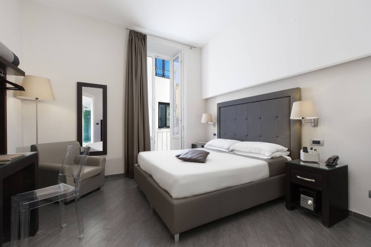 A modern hotel room with a large bed, a desk, a chair, and a nightstand with a lamp.