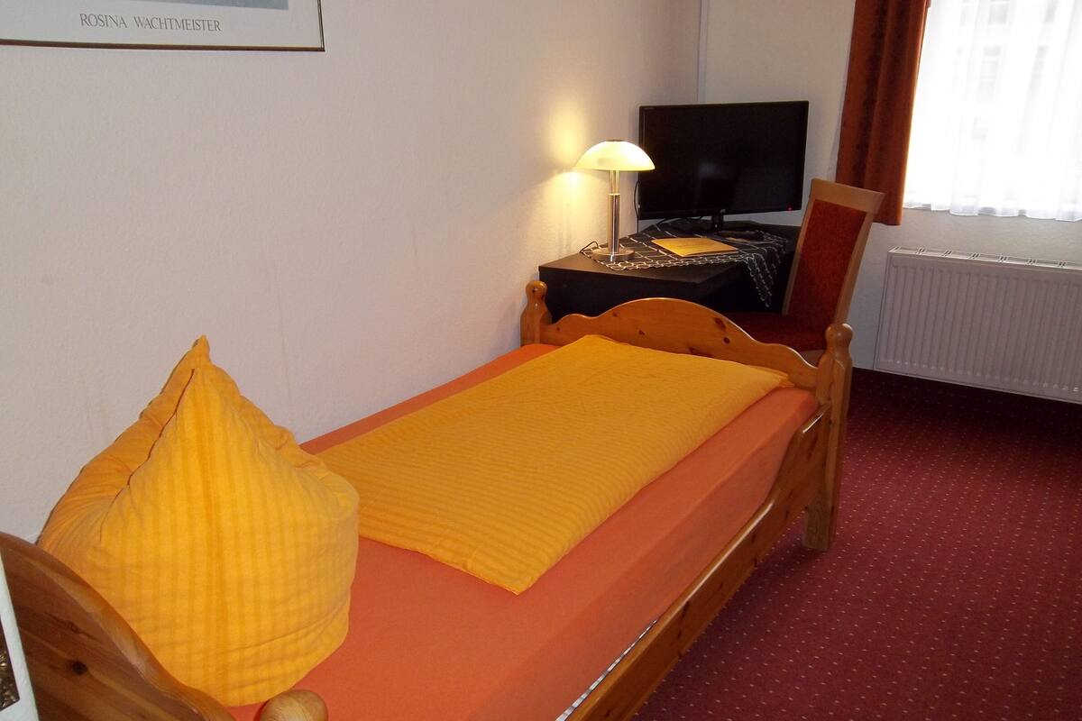 A single bed with a wooden frame, a yellow blanket, and a matching pillow. There is a small bedside table with a lamp and a television. The room has a red carpet and a white radiator.