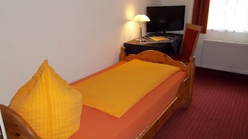 Una cama individual con estructura de madera, un almohadón amarillo y una manta. Hay un televisor sobre una mesita de noche.