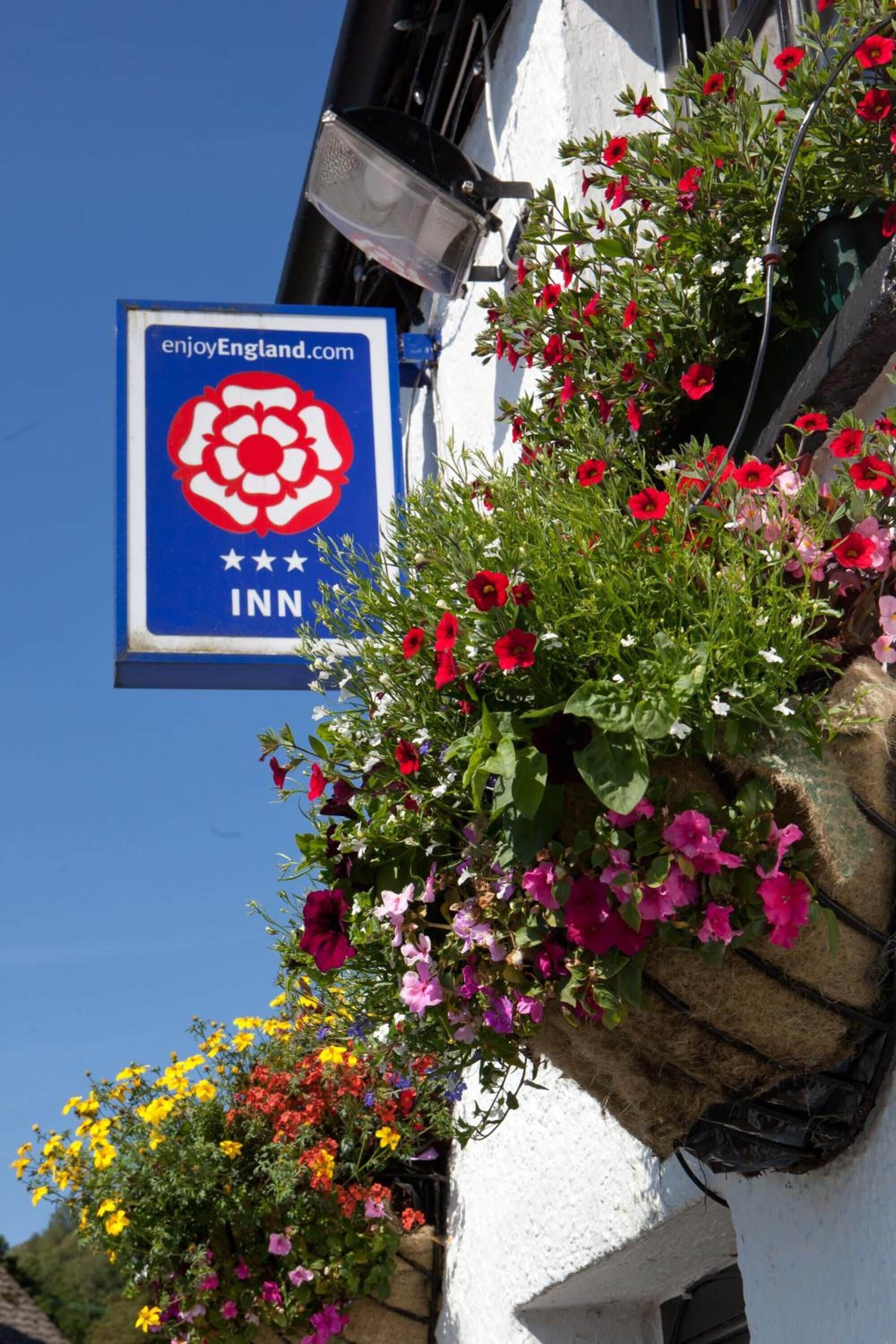 Um edifício com uma cesta suspensa cheia de flores vermelhas, cor-de-rosa e amarelas. Há uma placa com o logotipo de Enjoy England e o texto 